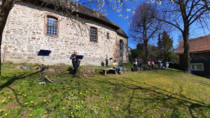 Ein Mann steht vor einem alten Steinhaus auf einem grasbewachsenen Hügel, während andere Menschen im Hintergrund sitzen.