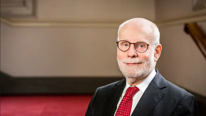 Ein älterer Mann mit Brille und Krawatte posiert vor einem roten Vorhang.