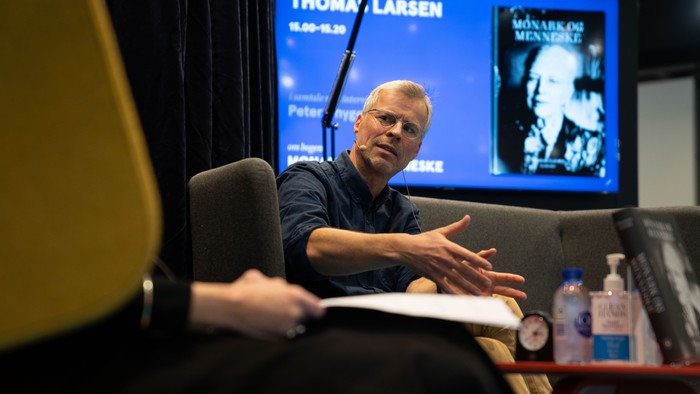 En mand sitter på en sofa og taler til en person, mens der vises et interview med Thomas Larsen på skærmen bagved.