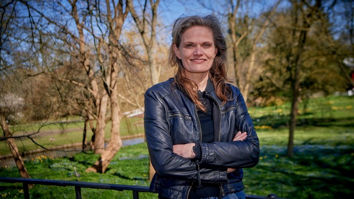 En kvinde med langt hår står med armene krydsede i en park.