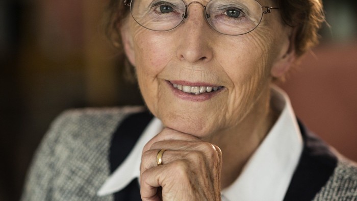 En eldre kvinde med briller og kort hår, smiler og har hovedet på hånd.