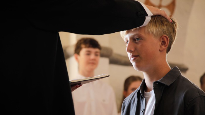 En ung dreng med blond hår står med en hånd på sit hoved og ser til siden.