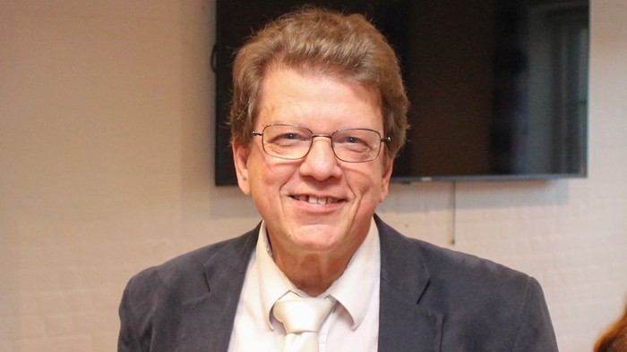 En smilende mand i anzug og briller står foran en blå stol.