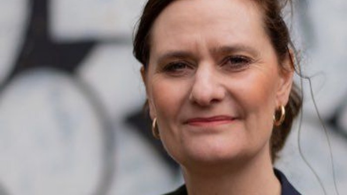 En kvinde med kort, mørkt hår og en blå jakke står foran et murværk med klokker.