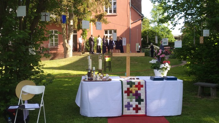 Im Garten vor einem Haus stehen Tische mit Blumen und eine rote Teppichrolle, während Menschen im Hintergrund stehen.