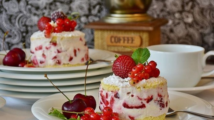 Zwei Desserts mit Erdbeeren und Kirschen auf Tellern, Kaffeetasse und Kaffeemühle im Hintergrund