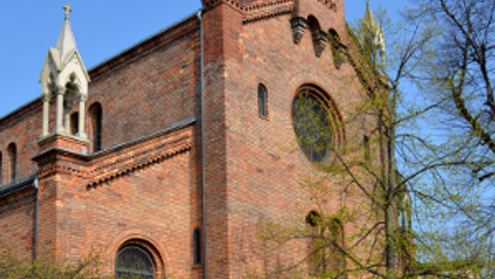 Ein rotes Backsteingebäude mit zwei Türmen und einem Kreuz darauf, umgeben von Bäumen und einem klaren blauen Himmel.