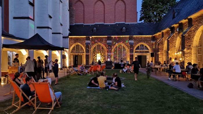 Im Innenhof eines Gebäudes sitzen Menschen auf Liegestühlen und Tischen, genießen Getränke und Gespräche bei Dämmerung.