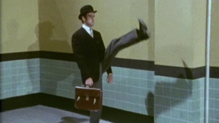 Mand i mørkt kostume og hat holder en brune mappe og springer i et rum med blå og gule vægfliser.