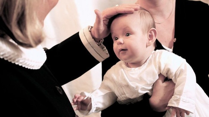 En baby i hvidt tøj bliver holdt af en person i sort og hvid klædning.