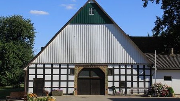 Weißes Holzhaus mit grünem Dach und schwarzer Tür, umgeben von Bäumen und Blumen