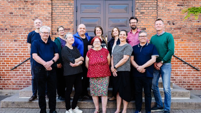 En gruppe af 13 personer står sammen foran en rød mur og en dør.