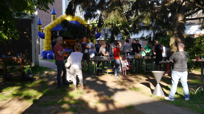 Eine Gruppe von Menschen feiert im Freien, einige stehen, andere sitzen an Tischen, ein bunter Bouncy Castle ist im Hintergrund.