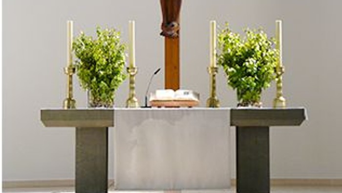 Ein hölzernes Kruzifix steht auf einem Altar mit zwei Kerzenhaltern und Topfpflanzen in einer Kirche.