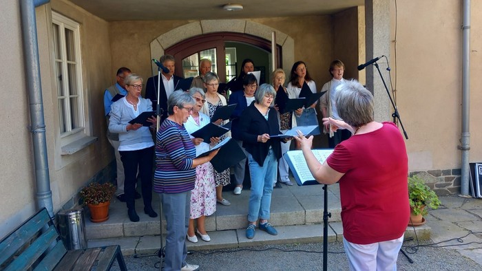 Ein Chor probt vor einem Haus, eine Frau dirigiert.