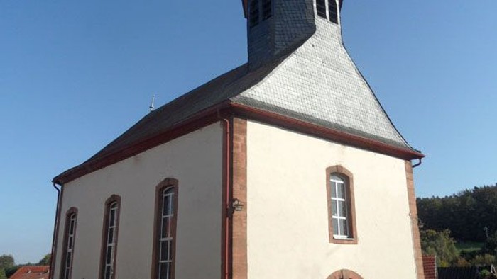 Kleine weiße Kirche mit roter Umrandung und Kirchturmspitze, vor einem klaren blauen Himmel.
