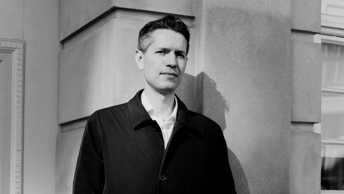 Mand i mørk jakke og hvid skjorte står ved murvæg.