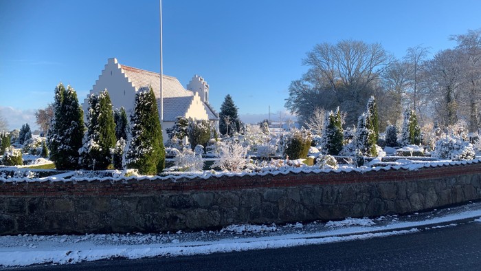 En snedækket kirkegård med en kirke i baggrunden og en mur foran.
