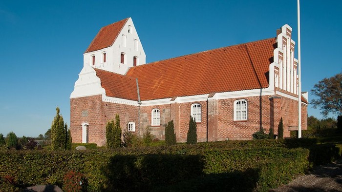 En rød og hvid kirke med spiret og et tårn, omgivet af grønne buske og en klar blå himmel.
