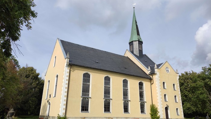 Gelbe Kirche mit hohem Turm und grünem Dach, umgeben von Bäumen und Gräbern