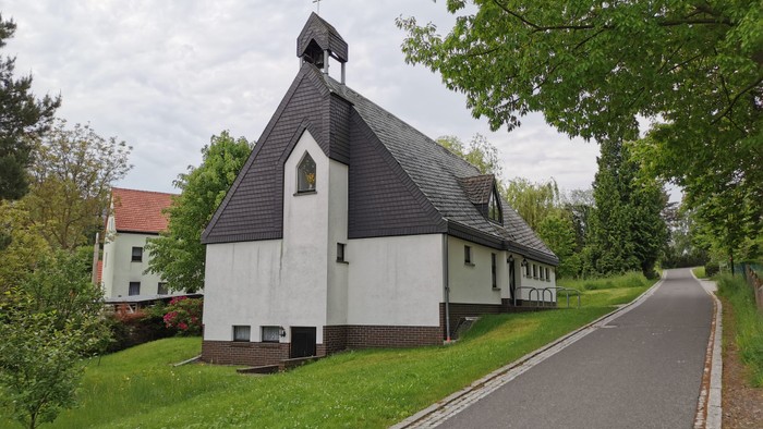 Kleine Kirche mit schwarzem Dach und weißer Fassade, umgeben von Bäumen und einem Weg.