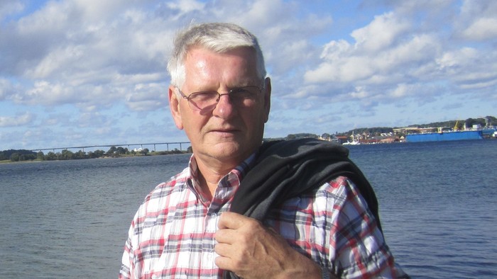 En mand med briller og rød-hvid fløjel stander ved vandet med en kamera.