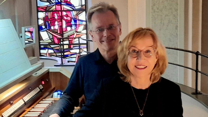 Ein Mann und eine Frau sitzen an einem Klavier, vor einem bunten Buntglasfenster.