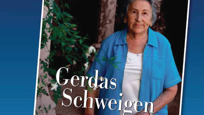 Eine ältere Frau in blauer Jacke steht vor einem Baum und einem Gebäude.