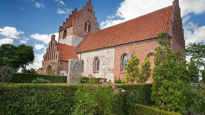 Gammel kirke med røde tagtegnere og omgivelser af grønne buske og træer