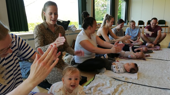 En gruppe af voksne og børn sidder på gulvmatte i et rum med store vinduer.