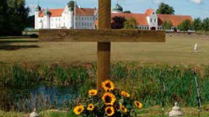 En grav med kors, blomster og lys i forgrunden, og et slot i baggrunden