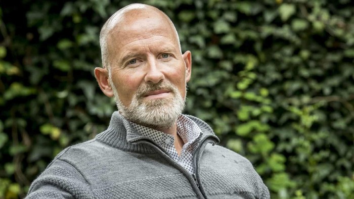 En bald, bart mands ansigt, der ser direkte i kameraet med en grøn, bladrig bakgrund