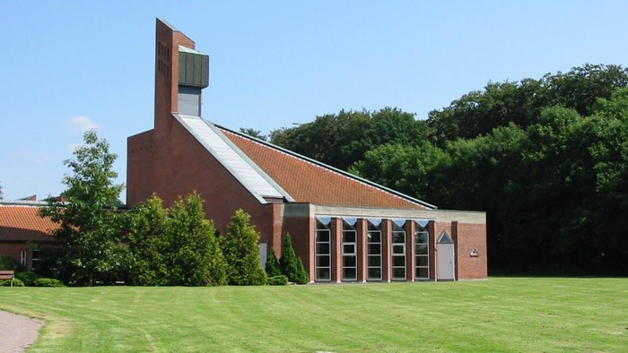 Et moderne kirkebygning i rød tegl med skråt tårn og store vinduer til en grøn og åben plads.