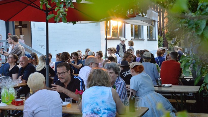 "Overfyldt udendørs spisested med mennesker, der sidder ved borde, nogle med drikkevarer og mad, under parasoller og træer."