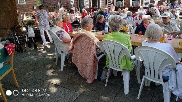 Eine Gruppe älterer Menschen sitzt im Freien an Tischen und genießt ein Essen.