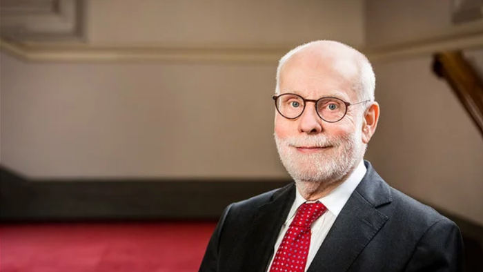 Ein älterer Mann mit Brille und Krawatte posiert vor einem roten Vorhang.