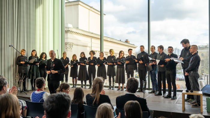 Ein Chor tritt vor einem Publikum auf, geleitet von einem Dirigenten auf einer Bühne.