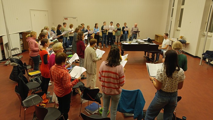 Eine Gruppe von Menschen proben in einem Raum Musik, einige stehen, andere sitzen und spielen Instrumente.