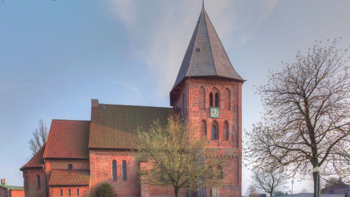 Ein rotes Backsteingebäude mit hohem Turm und spitzen Dach, umgeben von Bäumen und grünem Gras unter einem klaren blauen Himmel