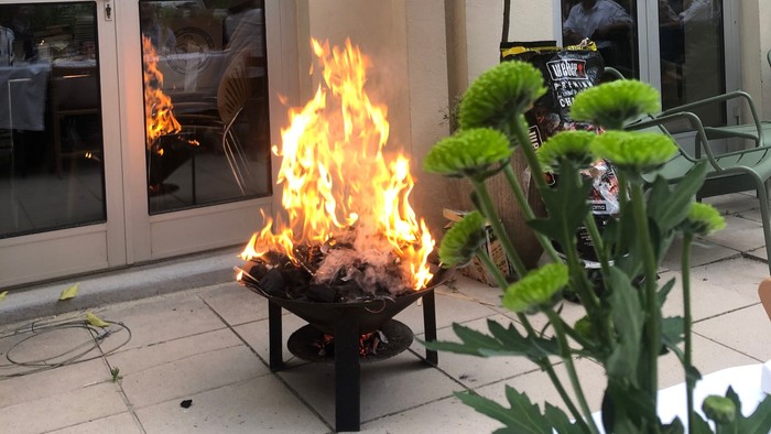 En lille ild i en firebåge på en terrasse foran et café med grønne planter og store vinduer
