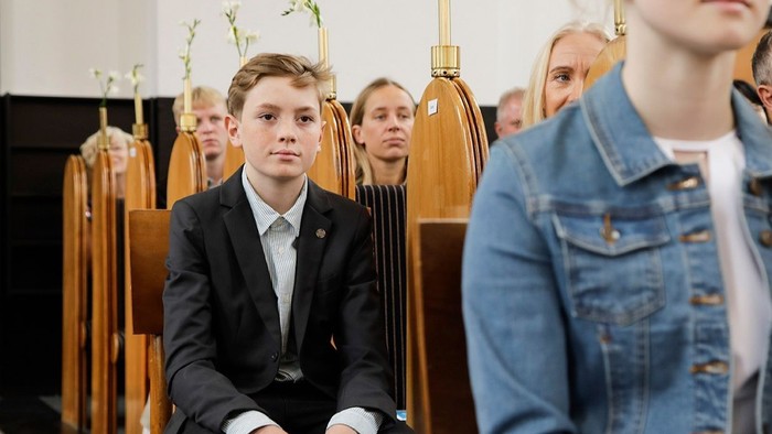 En ung dreng i formel klædning sidder mellem to voksne, mens en pige i blå jeanjacke ser på ham.