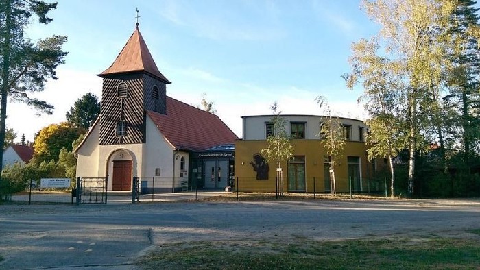 Kleine Kirche mit rotem Dach neben einem größeren gelben Gebäude auf einem Parkplatz.