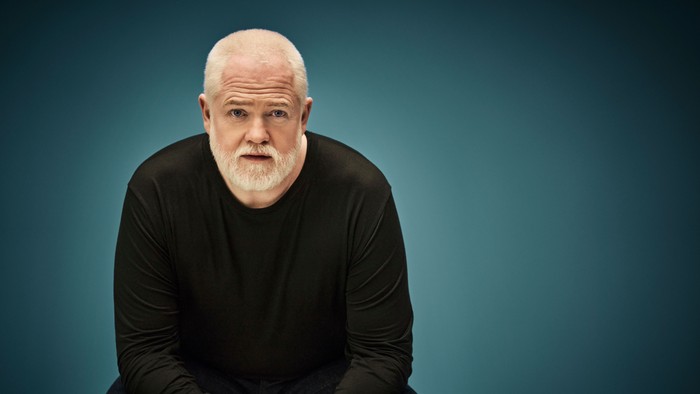 En bald, bart mands sidder med henderne samlet og ser tæt på kameraet.