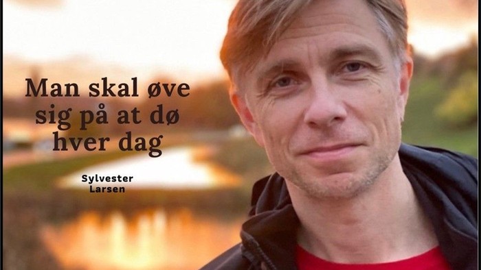 En mand med blå øjne og kort hår, der ser direkte i kameraet med en sø og skov i baggrunden.