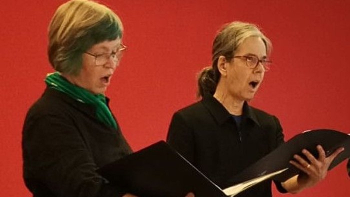 Zwei Frauen singen aus Notenblättern, beide tragen Brillen und dunkle Kleidung.