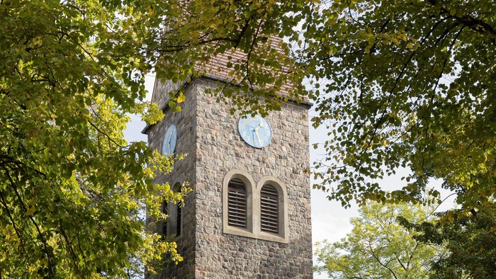 Der Kirchturm aus Stein mit Uhr und Fenster steht in einem Park.