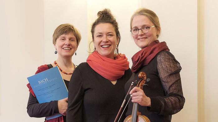Drei Frauen posieren, eine hält ein Buch, eine eine Geige