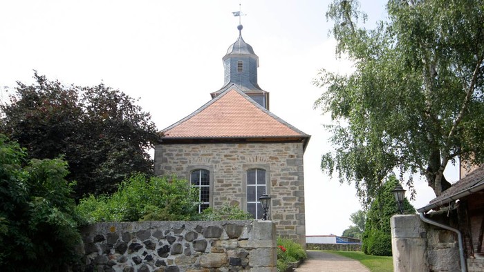 Kleines Steingebäude mit Wetterhahn, umgeben von Grünflächen.