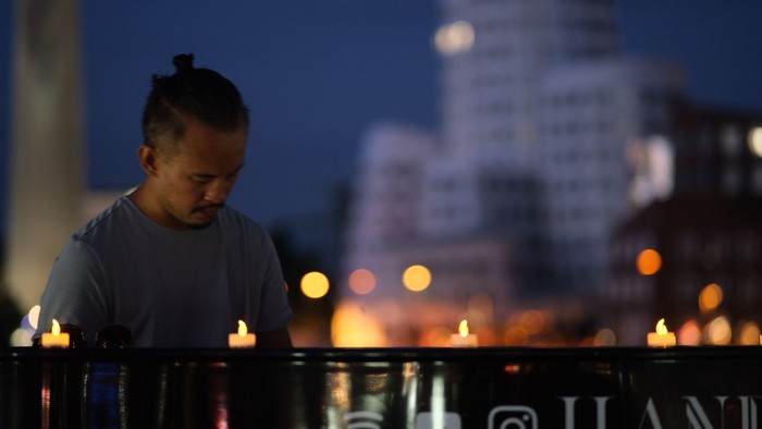Ein Mann beugt sich über ein Geländer mit brennenden Kerzen in der Nacht, im Hintergrund eine Stadtlandschaft.