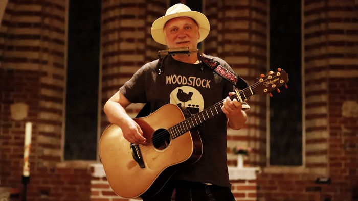 Ein Mann in einem Kirchenschiff spielt Gitarre und trägt einen Strohhut.